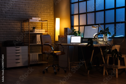 Interior of dark office with workplace, shelf unit and glowing lamps at night