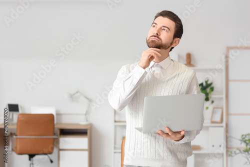 Thoughtful young man with laptop looking upwards in office