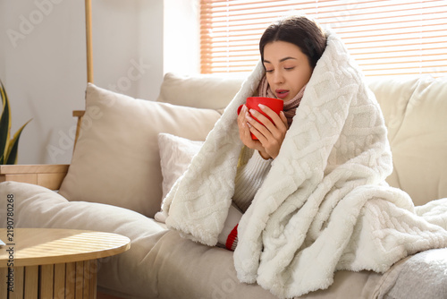 Frozen young woman with warm plaid and cup of hot tea sitting at home on wint...