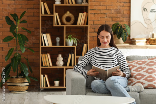 Young woman sitting on sofa and reading book in living room