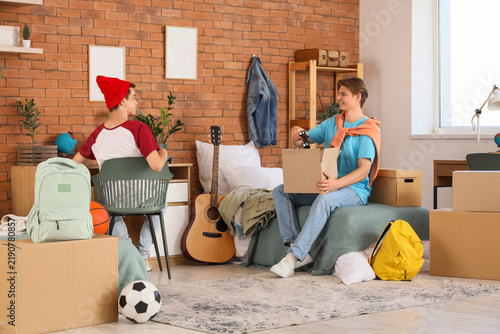 Male student taking game pad out of box and his roommate in dorm room on movi...