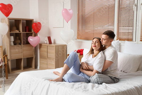 Beautiful young couple sitting on bed and celebrating Valentines Day at home