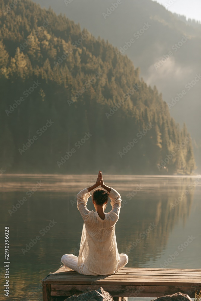 Fototapeta premium Woman doing yoga pose on a lakeside pier with mountain reflections for mindfulness background and wellness banner