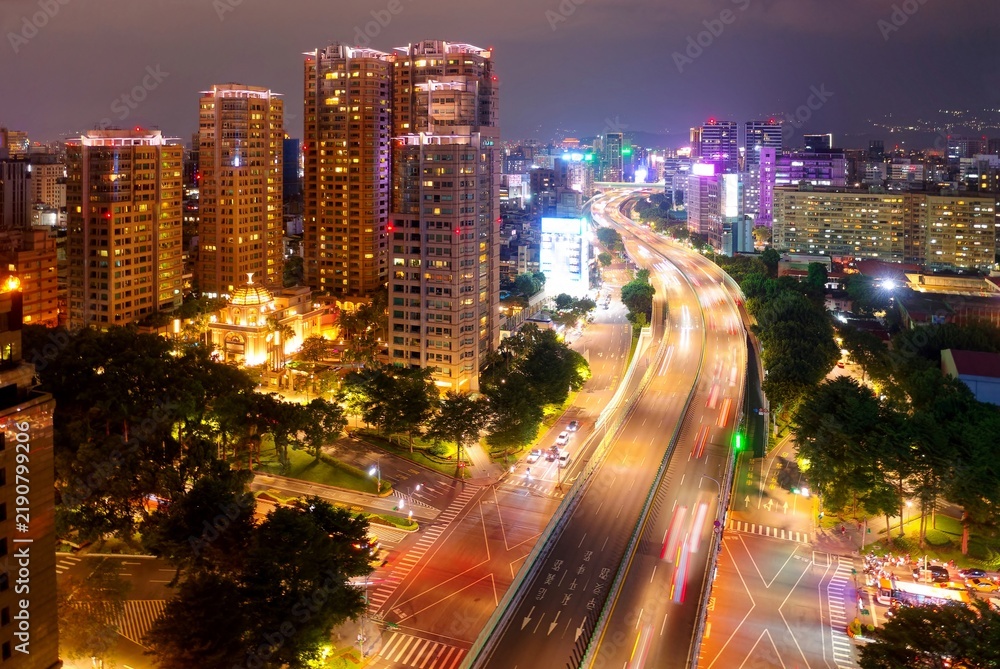 Fototapeta premium Aerial view of Jianguo S. Rd Expressway flying over Ren'ai Road, in Taipei, Taiwan, with residential towers of luxury apartments standing by the wooded avenue and city lights dazzling in the night