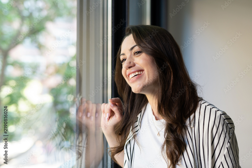 Obraz premium Young woman with a radiant smile standing by a window, looking out with hopeful, relaxed optimism and peaceful contemplation, enjoying natural light and a bright mood