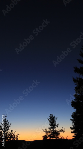 Vertical view of a tranquil twilight landscape in Quebec, Canada, with a vast deep blue sky fading into a warm orange horizon over tree silhouettes.