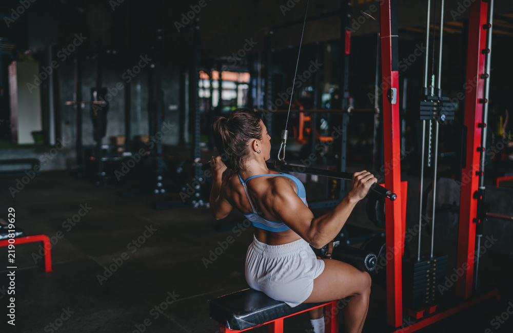 Fototapeta premium Three quarter back view of Hispanic woman doing lat pulldown exercise on machine, athletic female training upper body back muscles in gym setting, strength workout, copy space.