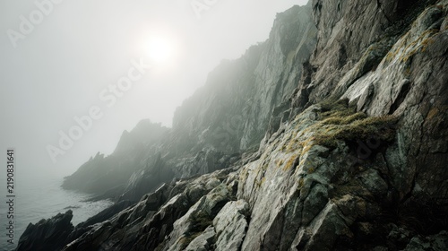 Misty Coastal Cliffs: Rugged, moss-covered cliffs meet the ocean under a mysterious, misty sky, with the sun faintly visible through the haze, creating a dramatic and serene landscape.