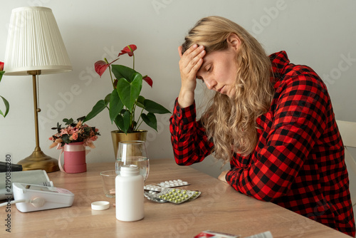 Exhausted sick woman holding her forehead while sitting near medicines, water and blood pressure monitor at home. Concept of headache, fatigue, illness symptoms and difficult recovery.