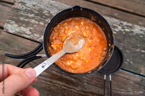 Obraz na płótnie Man stirring boiling camp stew with beans tuna and melted feta cheese using a spoon in a outdoor pot on wood