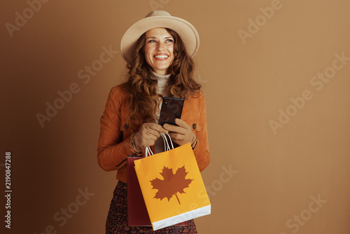 An elegant woman with wavy brown hair and a wide-brimmed hat smiles, holding her smartphone and colorful shopping bags featuring a distinct maple leaf design.
