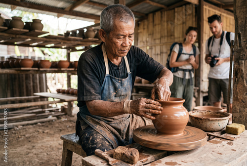 Senior artisan shaping clay pot on wheel as tourists observe in traditional rural pottery workshop