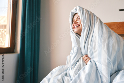 Happy young woman wrapped in a cozy blanket laughs in bed by the window at home, morning mood, comfort and relaxation concept.