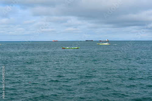 Fishing boats floating on calm ocean water with overcast sky and cargo ships on horizon	
