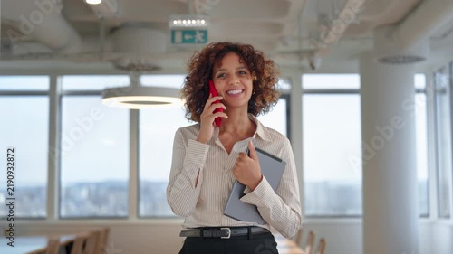 Latin business woman walking in coworking office talking on smartphone phone and holding digital tablet. Young hispanic teacher or corporate trainer makes client call near presentation board