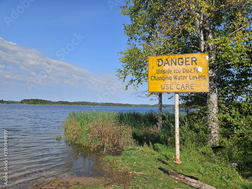 Obraz na plátně Humorous yellow sign warning of the dangers of thin ice  is in contradiction to the lake of calm water it is standing next to during the summer