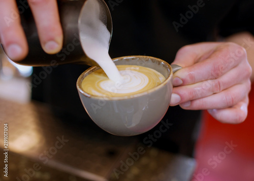 Latte art. Pouring foamed milk. Barista hand adding frothing creamy component from pitcher to beverage garnished coffee in ceramic cup.