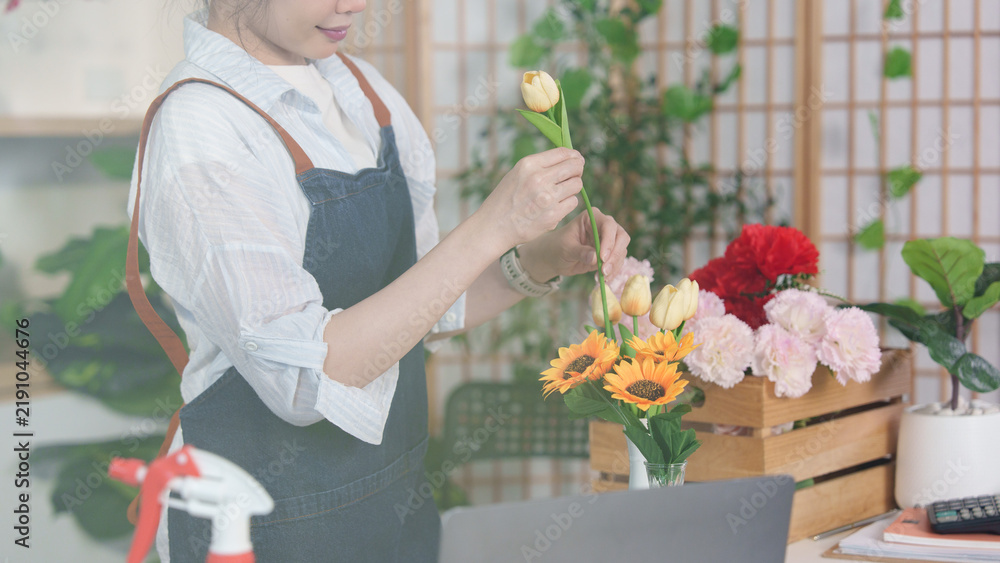 Fototapeta premium Asian female florist arranging yellow tulips in vase at flower shop counter for floral design