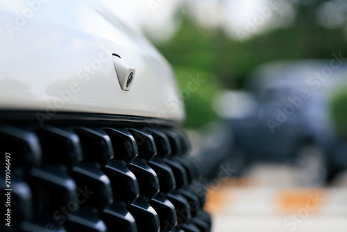 front facing camera mounted on white vehicle hood above textured black front grille, automotive exterior, angular parking assist camera lens used for driver assistance, surround view, parking safety