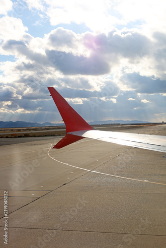 View from an airplane window showing a red winglet on the tarmac with a cloudy sky in the background.
