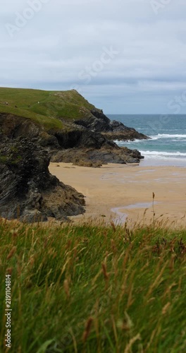 Sandy beach and rugged cliffs in Cornwall, England, UK, with breaking waves and coastal grass beneath a cloudy afternoon sky. Vertical handheld footage.