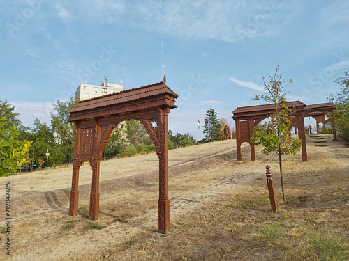 Foto From the Székely gates standing on the Vértói Hill in Szeged, two of them can be seen in sunny weather, with dried grass, leafy trees, and behind them, a multi-story residential building