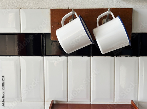 Two white vintage enamel mugs with blue rims hanging on wooden hooks against a white and black tiled kitchen wall. Perfect for retro cafe interior concepts.