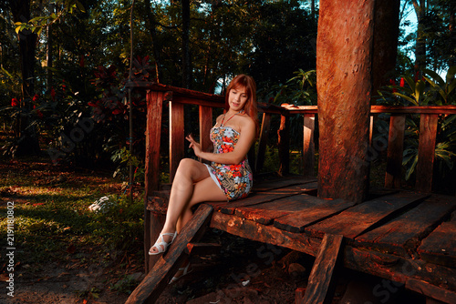 Fotografie A beautiful young Latina with reddish-brown hair sits cross-legged on a wooden kiosk floor, her flowery dress glowing in sunset light