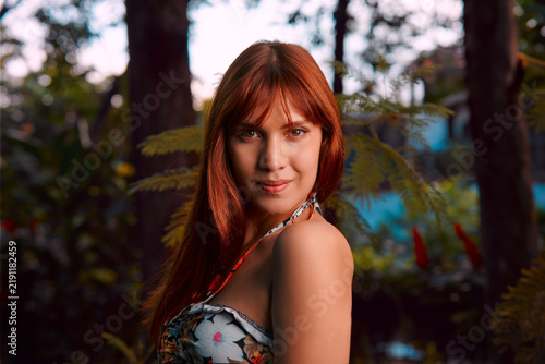 A close-up of a beautiful young Latina with reddish-brown hair in a flowery dress at a park, her face glowing in sunset light.