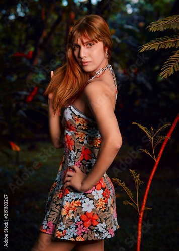 A beautiful young Latina with reddish-brown hair stands in an elegant, playful pose, her flowery dress glowing in sunset light, captured in wide angle.