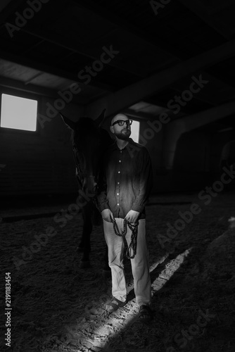Foto Dramatic black and white portrait of a man standing beside a horse inside a dark stable, with strong directional light, deep shadows and a cinematic atmosphere