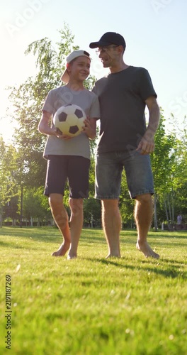Father and son walking in a park after a soccer game at sunset. Boy carries a football while they talk and smile. Vertical video showing family bonding and active lifestyle.