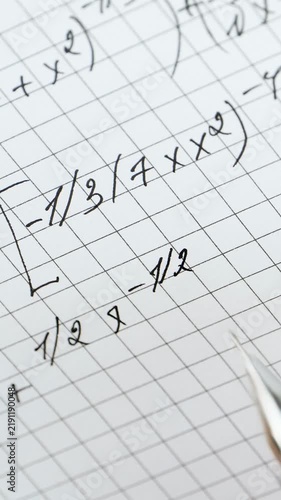 Close-up of mathematical formulas on graph paper. Student writes calculations with pen in classroom. Vertical shot captures learning atmosphere and academic focus.