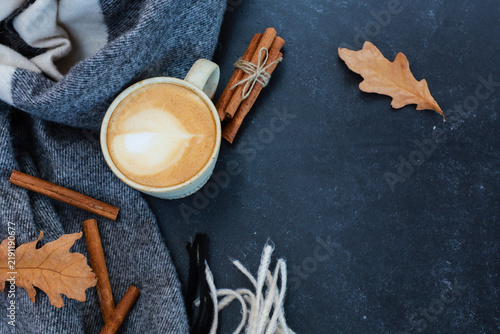 Cozy coffee beak in fall time. Cup of latte, cinnamon sticks, dry leaves on lack table. Top view with copy space. Autumn warming hot drink, calm cold morning vibe