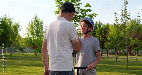 Father adjusts helmet for son as boy prepares to ride scooter in sunny park. Parent and child share safety lesson. Outdoor family moment promotes confidence, care, and active recreation.