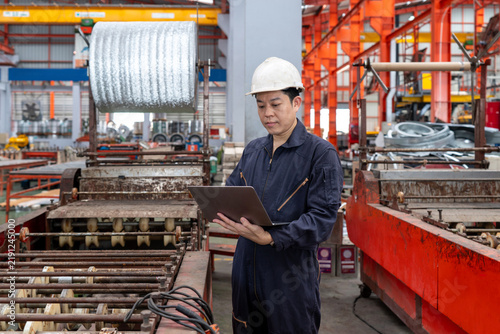 Принти на полотні A man in a hardhat and mechanic coverall dust protection suit stand in a busy warehouse, manage online document on laptop computer