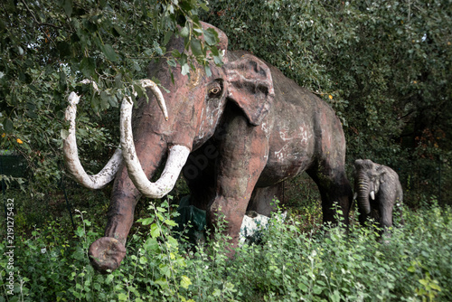 Riproduzione realistica a grandezza naturale di un mammut preistorico con grandi zanne ricurve e il suo cucciolo immersi nella fitta vegetazione verde di un parco tematico all'aperto.