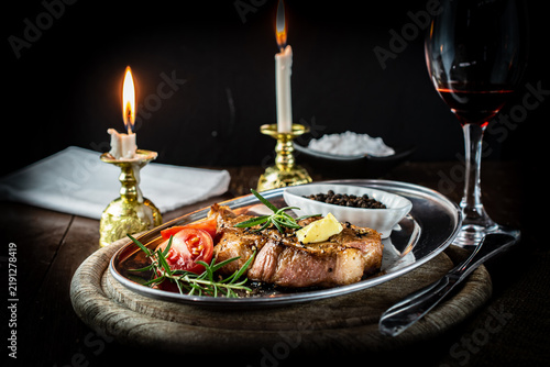 Romantic candlelit dinner featuring a perfectly grilled pork neck steak on a vintage metal platter, served with red wine, fresh rosemary, and ripe tomatoes in a warm, rustic atmosphere.