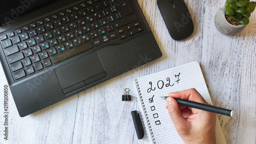 human hand writing goals for 2027 in notebook. Modern workspace with laptop, pen, computer mouse, and a succulent, idea planning for the new year, productivity, and organization.