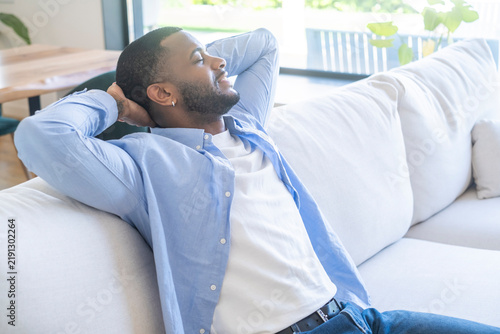 Photography Man relaxed on a couch in a bright living room, leaning back with hands behind his head, eyes closed and smiling, enjoying a peaceful moment of home comfort, leisure relaxation