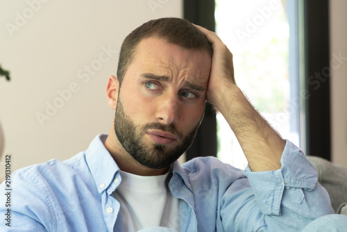 Fotografía Bearded man sitting on a sofa at home looking worried and pensive, resting his head on his hand while contemplating a problem, showing stress, concern and thoughtful emotion