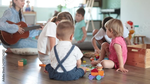Toys are scattered on floor, children are playing with cubes and cars, teacher is playing guitar. Girl babysitter accompanies children, twang guitar during childrens play with toys activities.