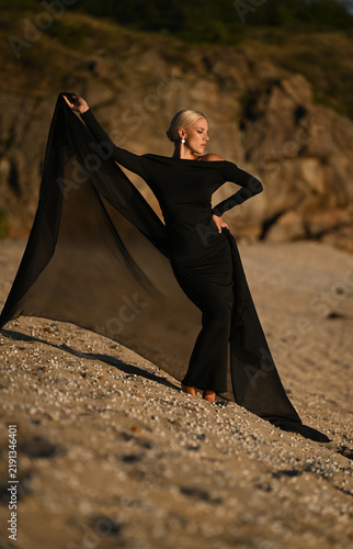 A beautiful blonde woman in a long black dress stands on the beach. Sunset.