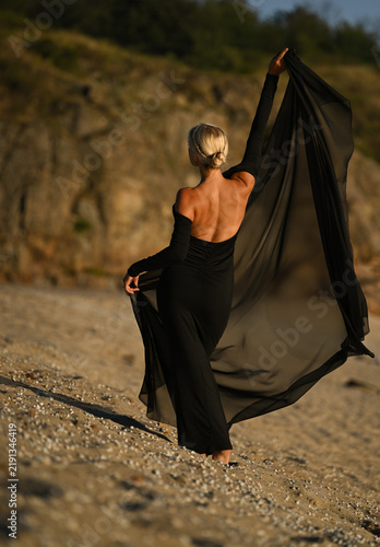 A beautiful blonde woman in a long black dress stands on the beach. Sunset.