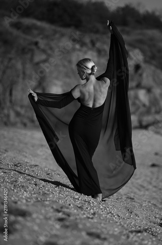 A beautiful blonde model in a long black dress stands on the beach. Sunset.