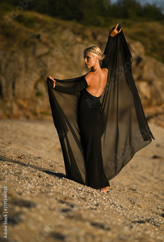 A beautiful blonde model in a long black dress stands on the beach. Sunset.