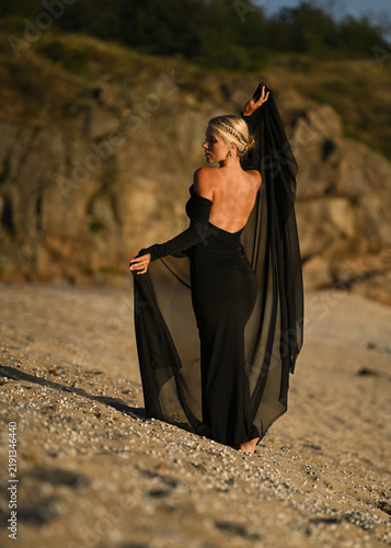 A beautiful blonde model in a long black dress stands on the beach. Sunset.
