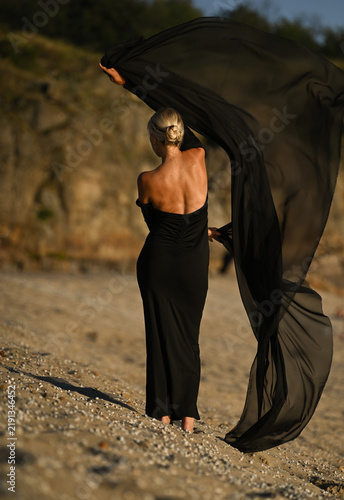 A beautiful blonde model in a long black dress stands on the beach. Sunset.