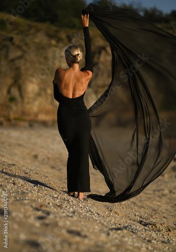 A beautiful blonde woman in a long black dress stands on the beach. Sunset.