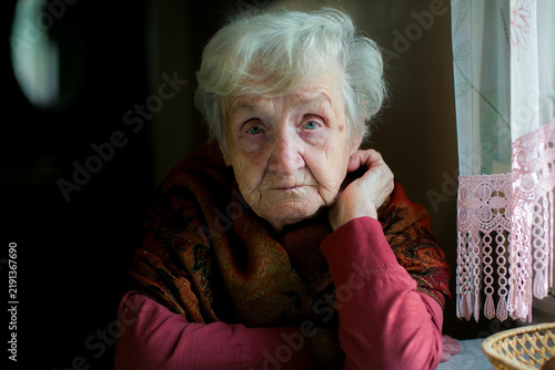 Close up portrait of an elderly woman sitting by a window with her hand beside her face and looking at the camera